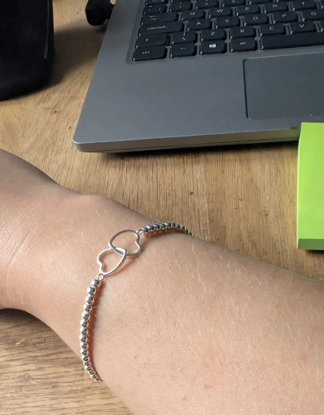 A silver beaded bracelet with two connected hearts on Ania’s wrist, resting on her desk beside her laptop.