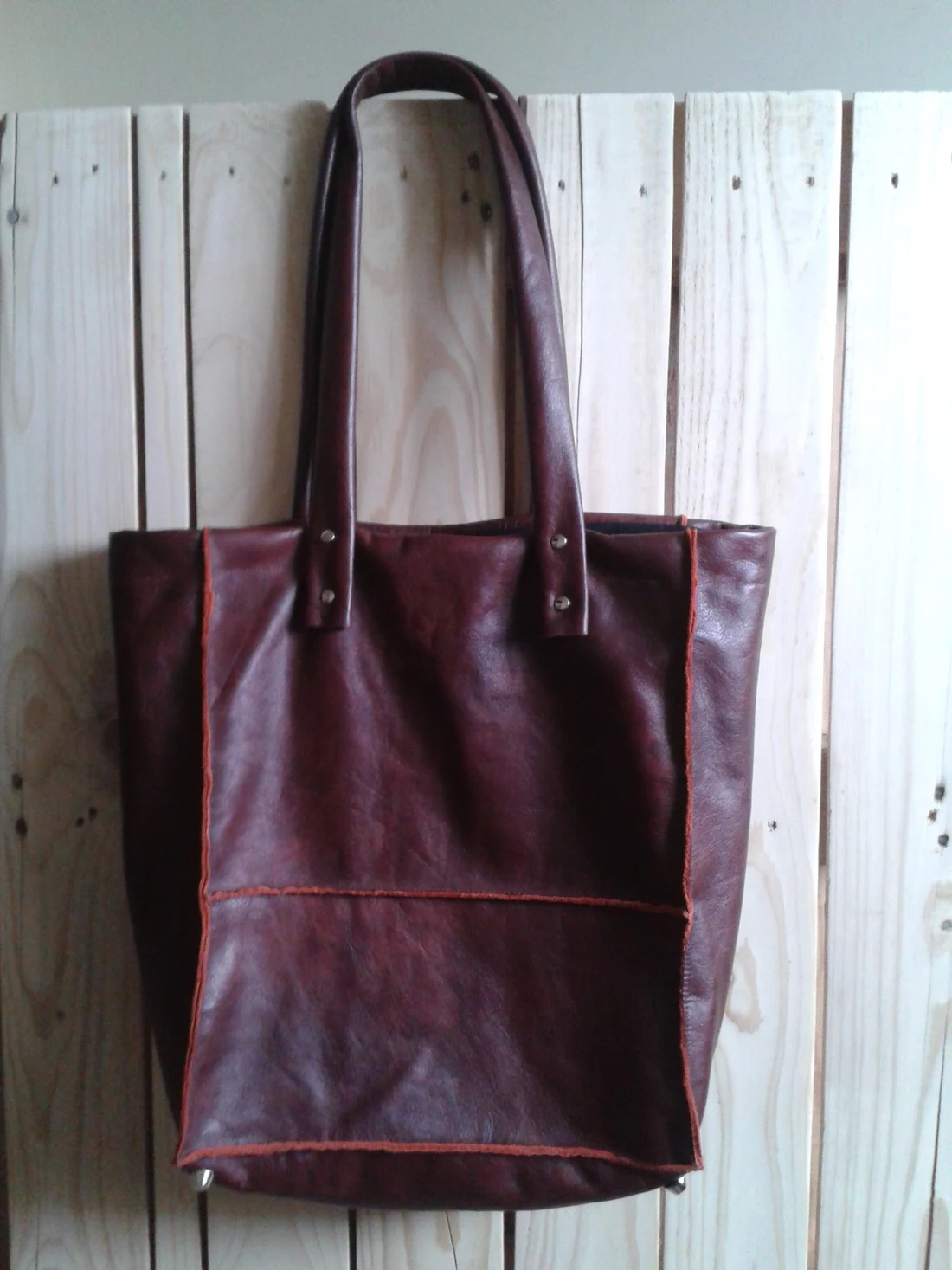 A handmade burgundy leather tote bag with long handles and hand stitched orange edging, hanging on pale wooden boards.