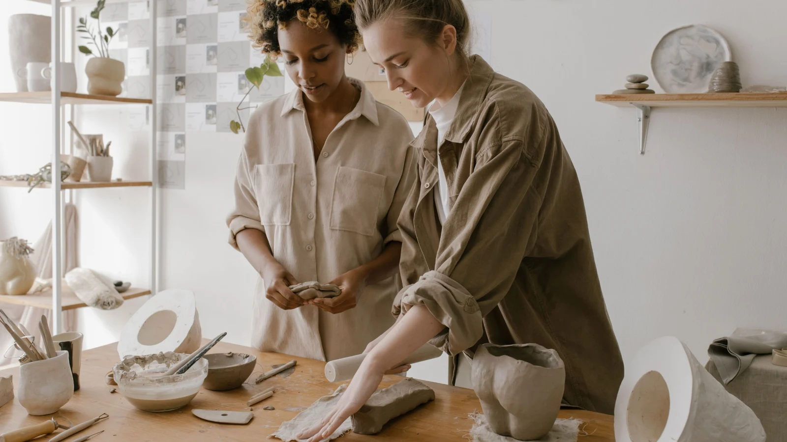 Two women working with clay together at a table in a pottery studio, surrounded by tools and finished pots.