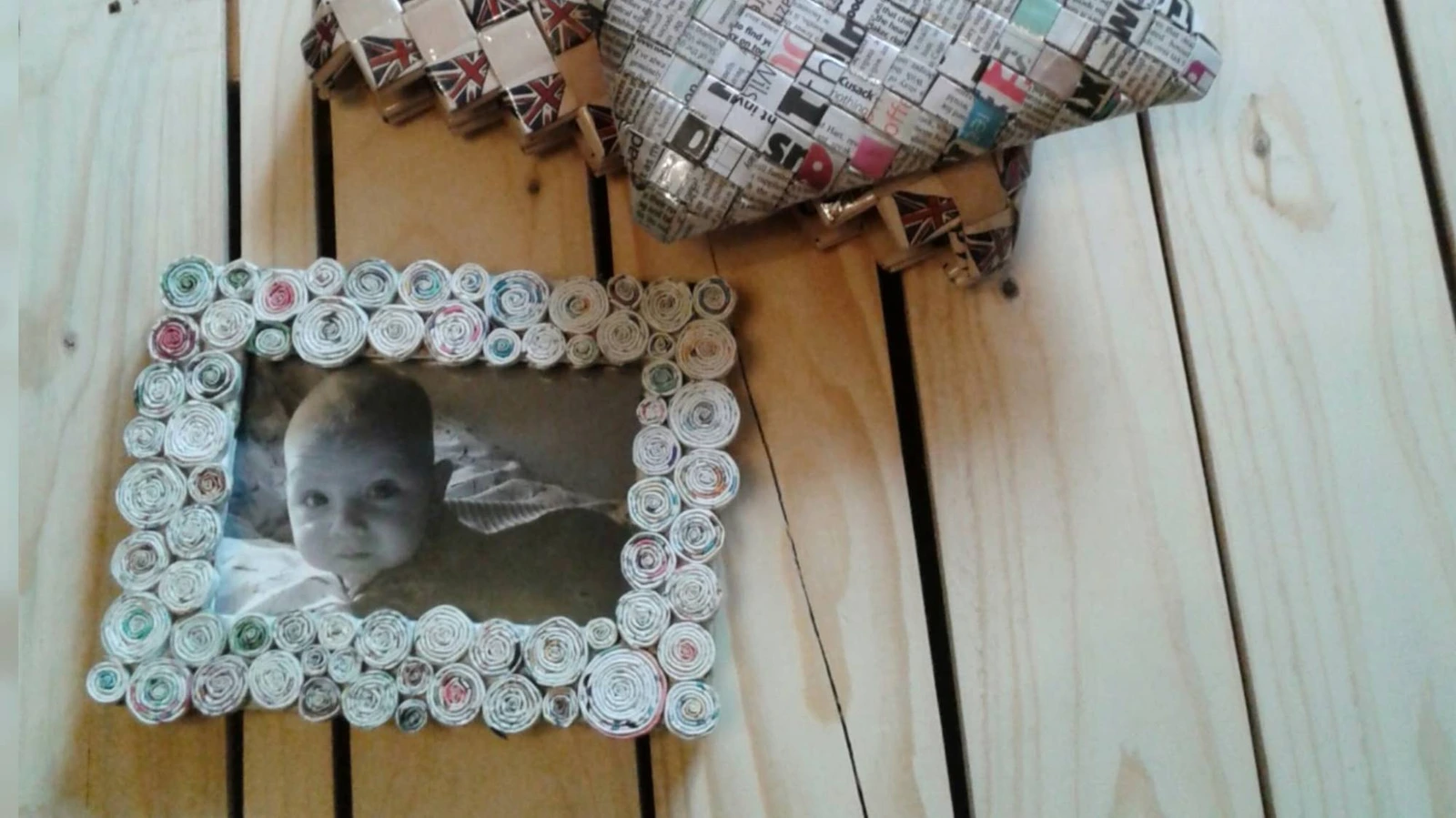 A handmade picture frame edged with tightly rolled paper spirals, holding a black and white photograph of a baby, on pale wooden boards.
