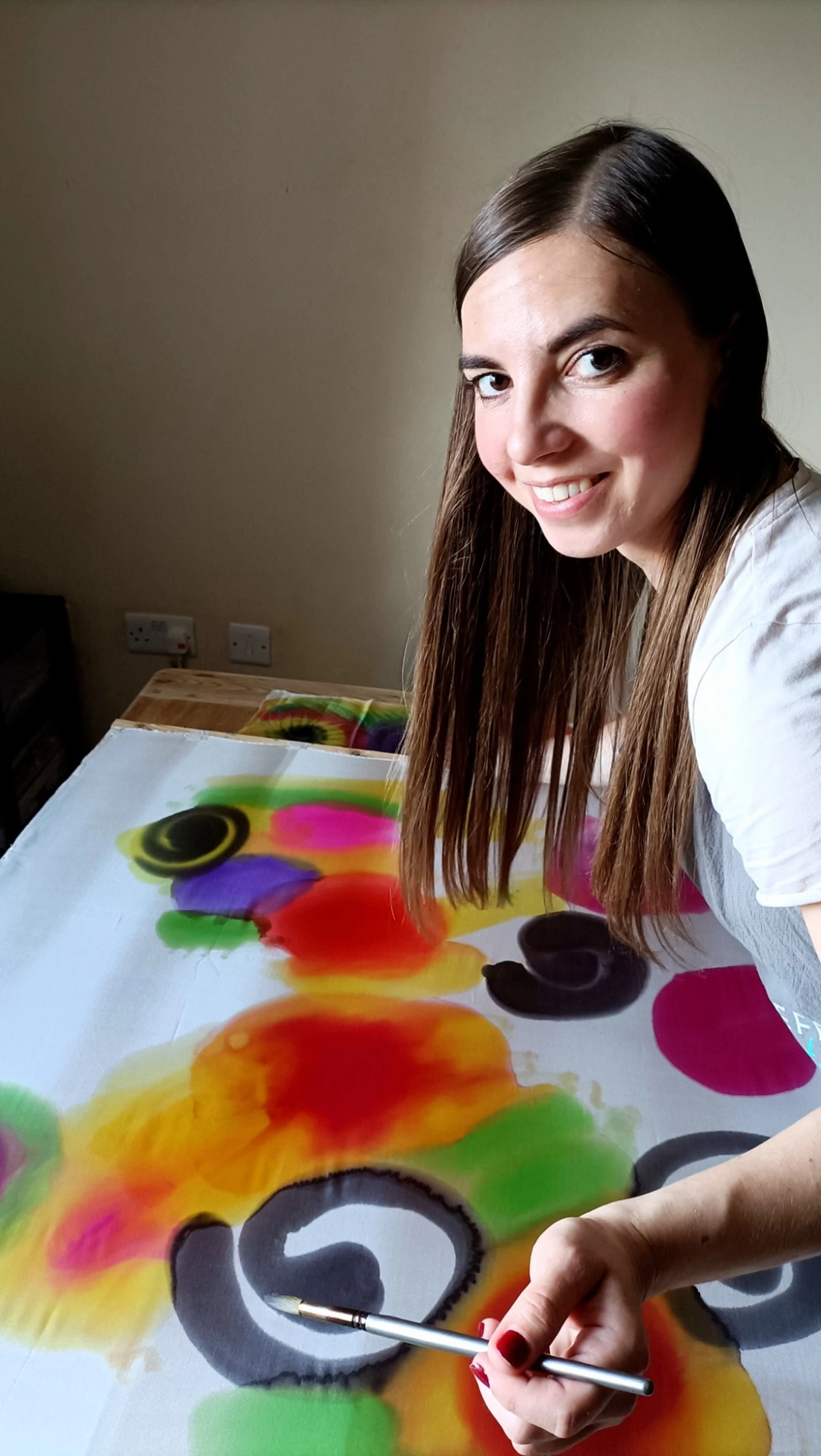 Ania smiling as she paints a large colourful piece of silk, holding her brush towards the camera.