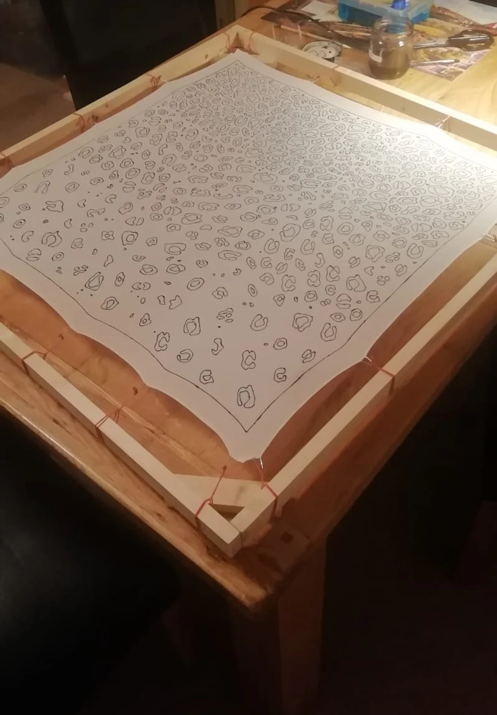 A large square of white silk stretched on a wooden frame on a table, its leopard pattern outlined in ink and waiting to be painted.