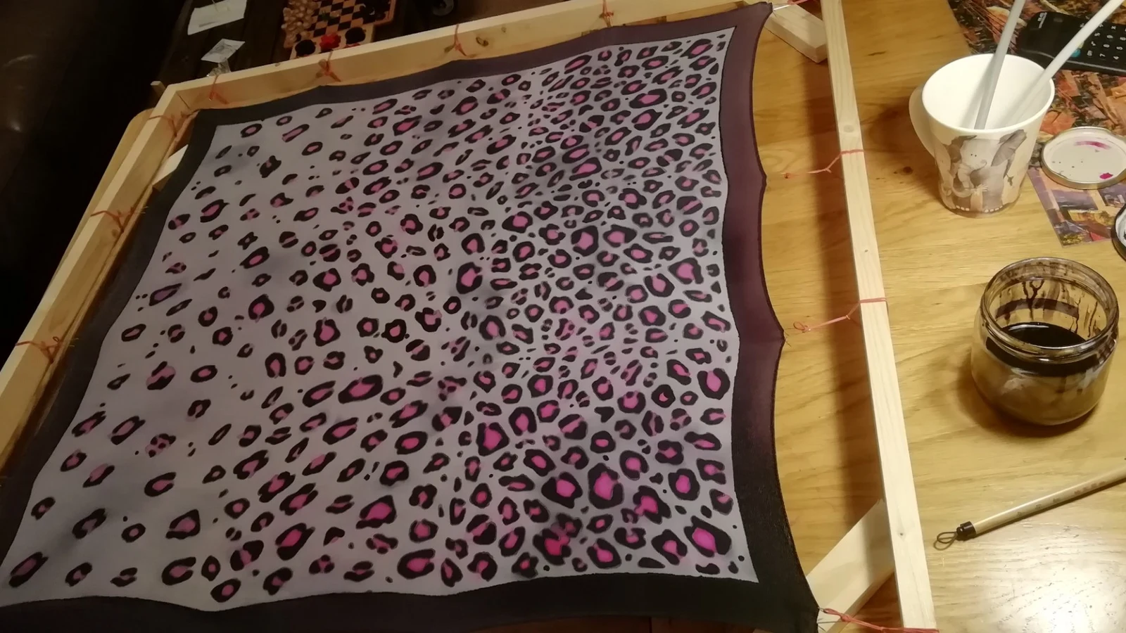 A silk scarf painted with pink and black leopard markings, stretched on a wooden frame on a table at home, with dye and brushes beside it.